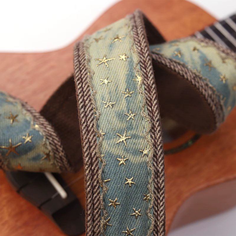 Sangle ukulélé en denim bleu délavé à étoiles dorées posée sur un ukulélé en acajou